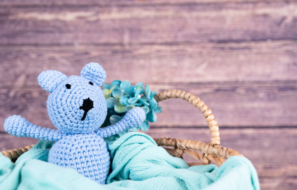 A blue crocheted bear, with a blue blanket, in a wooden basket.